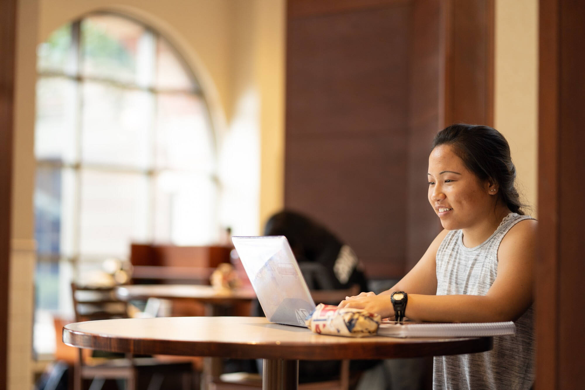 Student smiles while studying at laptop on round wooden table with notebooks and snacks, in warm-lit campus space with arched window and another student visible in blurred background.